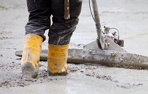 Eine Strasse wird betoniert. Der Beton wird von einer Person mit einer Schaufel verteilt.